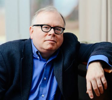Environmental portrait of a pianist in glasses and sport coat, in a chair near a window