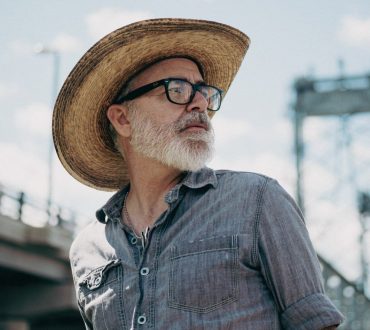 Environmental portrait of a bearded man in a straw hat and glasses against an overcast sky