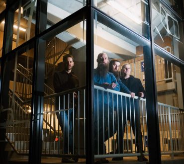 Band portrait of four men photographed through a glass stairwell at night