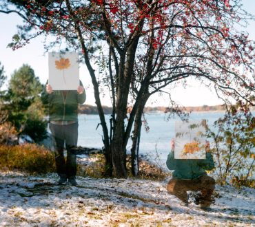 Two artists holding botanical prints in front of their faces at a snowy New England waterfront