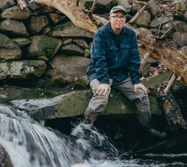 Environmental portrait of a man in a flannel shirt seated on rocks beside a rushing stream