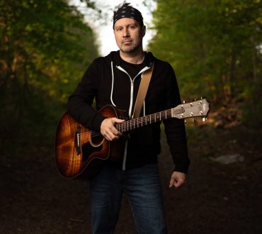 Outdoor portrait of a guitarist holding an acoustic guitar on a wooded path at golden hour
