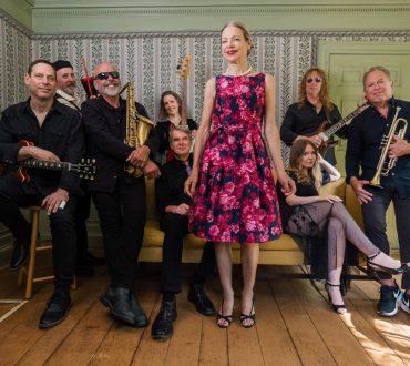 Band portrait of nine musicians in a vintage-wallpapered parlor, holding instruments
