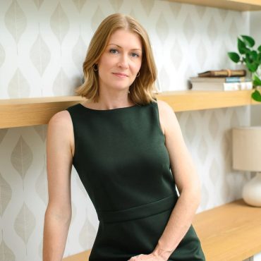 Environmental business portrait of a woman in a dark green dress leaning against a wood shelf