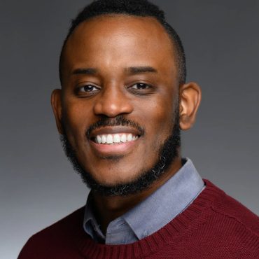Studio headshot of a smiling man in a burgundy sweater against a gray backdrop