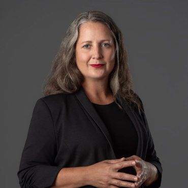 Studio headshot of a woman in a black blazer with hands folded against a gray backdrop