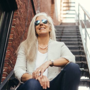 Outdoor environmental portrait of a woman in sunglasses seated on a fire escape in an urban brick setting.