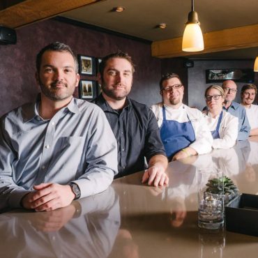 Team portrait of six restaurant staff seated at a bar, including chefs in white jackets