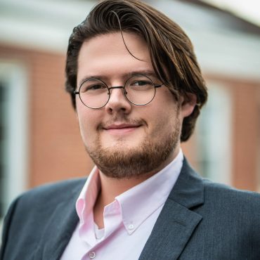 Outdoor business headshot of a young man in glasses and a gray blazer against a brick building
