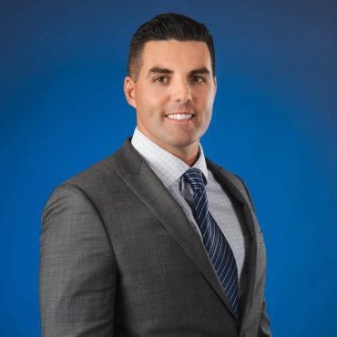 Studio headshot of a man in a gray suit and striped tie against a blue backdrop