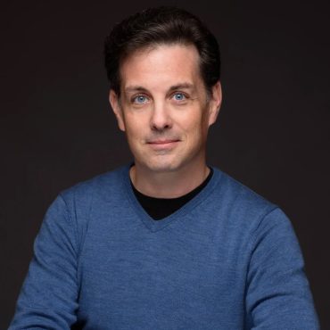 Studio headshot of a man in a blue sweater against a dark background