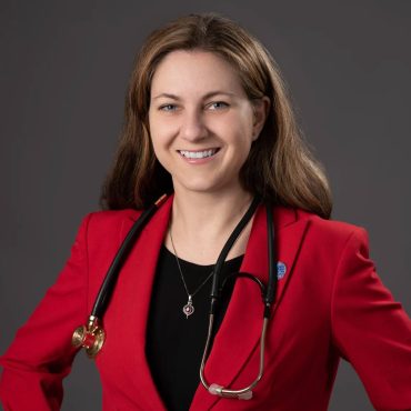 Studio headshot of a woman in a red blazer with a stethoscope against a gray backdrop