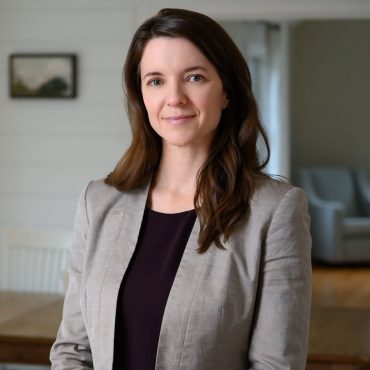 Environmental business portrait of a woman in a blazer standing in a softly lit home interior