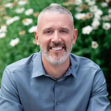 Outdoor natural light headshot of a man in a blue shirt against a garden backdrop