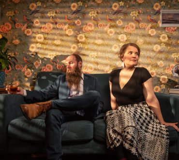 Environmental portrait of man and a woman musician sitting on a couch with warm venetian blind lighting.
